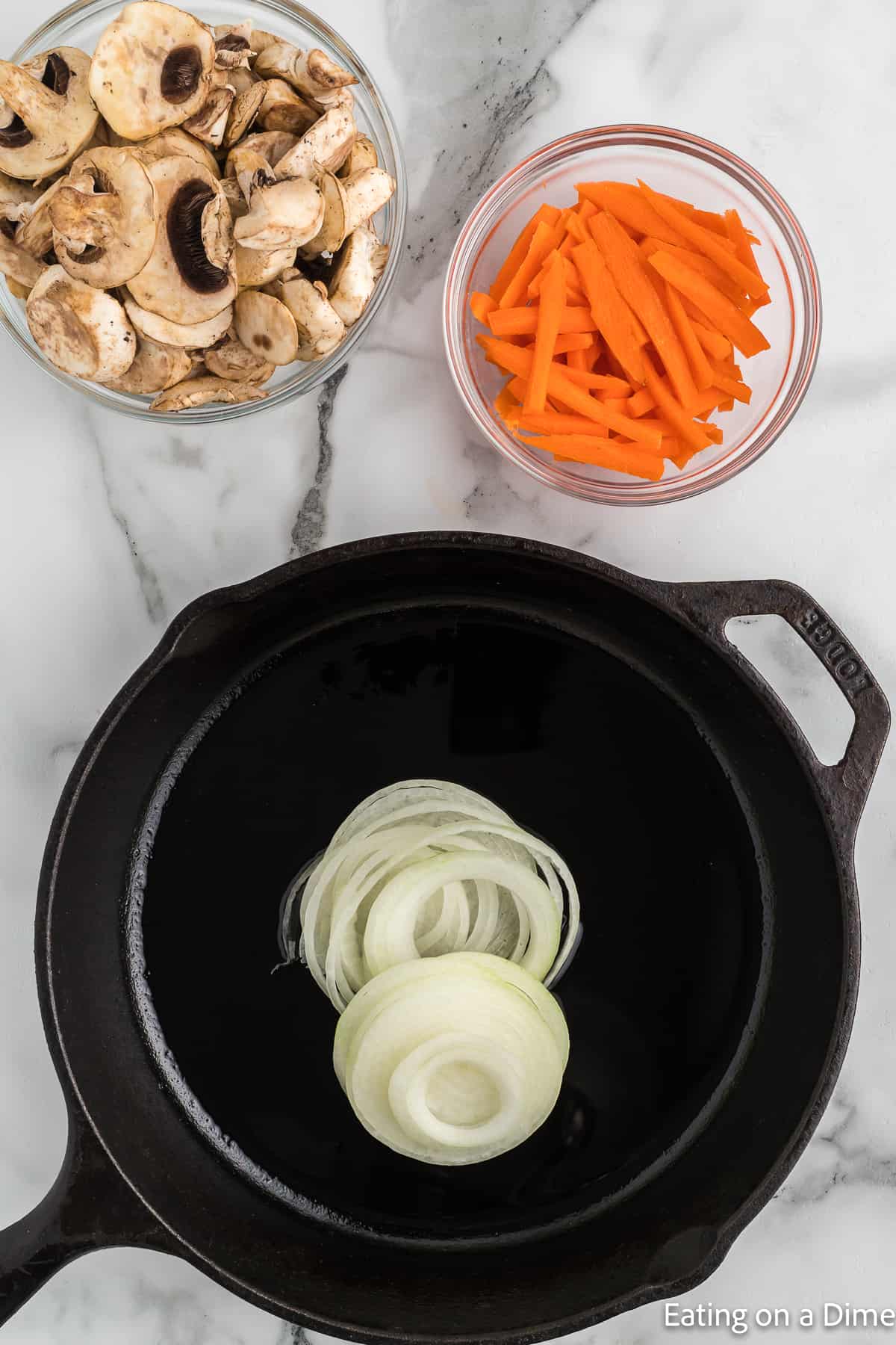 Cooking slice onions in a cast iron skillet with bowls of slice mushrooms and carrots