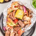 Surf and Turf Foil Packs filled with grilled shrimp, steak pieces, corn on the cob, red onion, tomatoes, and herbs, served on a black plate with a side of fresh parsley.