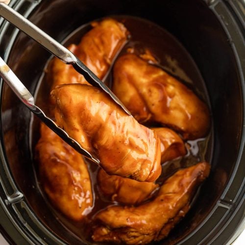 Slow Cooker Coca Cola Chicken breasts coated in a glossy brown sauce inside a slow cooker, with a pair of metal tongs lifting one piece out of the sauce.