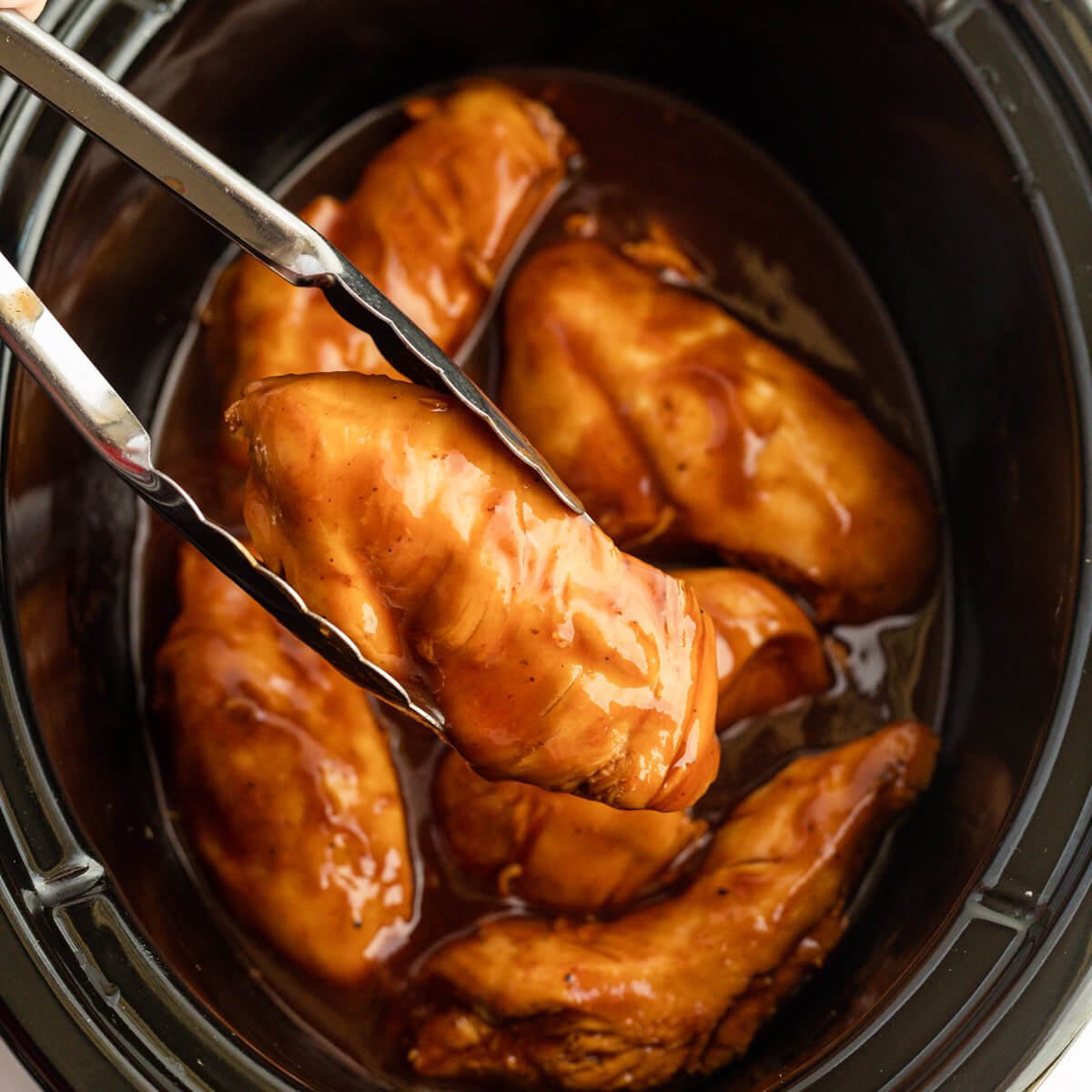 Cooked Coca Cola Chicken in the slow cooker with a piece on tongs