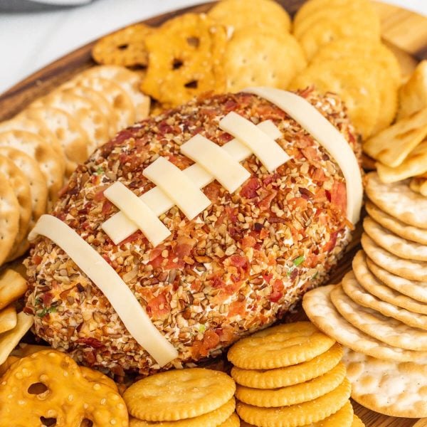 A Football Cheese Ball covered in chopped nuts and bacon, decorated with cheese strips to resemble football laces, sits on a wooden platter surrounded by assorted crackers—perfect for your next game day spread.