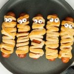 Five mummy hot dogs wrapped in crescent dough, each with candy eyes on top, are arranged in a row on a dark plate.