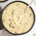 A bowl of creamy Zuppa Toscana Soup with chunks of potato, flecks of spinach, and a sprinkling of shredded cheese on top. A spoon rests in the bowl and a checkered cloth is nearby.