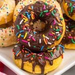 Two Cake Mix Donuts topped with chocolate glaze and colorful sprinkles are stacked on a plate, with more donuts covered in icing and sprinkles in the background.