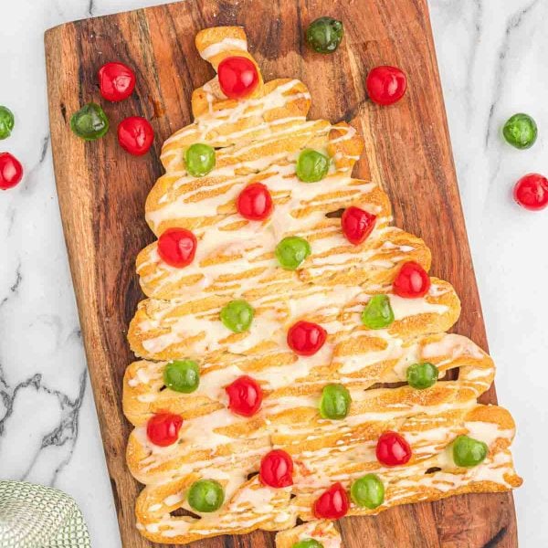 A Christmas Tree Coffee Cake pastry, drizzled with white icing and adorned with red and green candied cherries, sits on a wooden cutting board atop a marble surface.