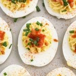 Close-up of Bacon Deviled Eggs topped with chopped chives, crispy bacon bits, and a sprinkle of paprika, arranged on a light-colored surface.