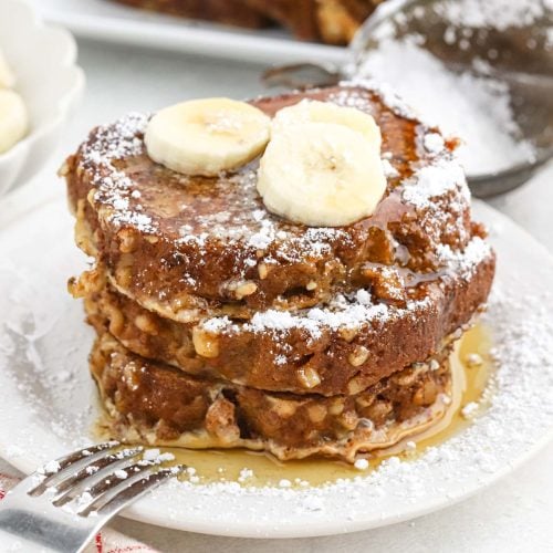 A stack of banana bread French toast topped with banana slices, dusted with powdered sugar, and drizzled with syrup, served on a white plate with a fork.