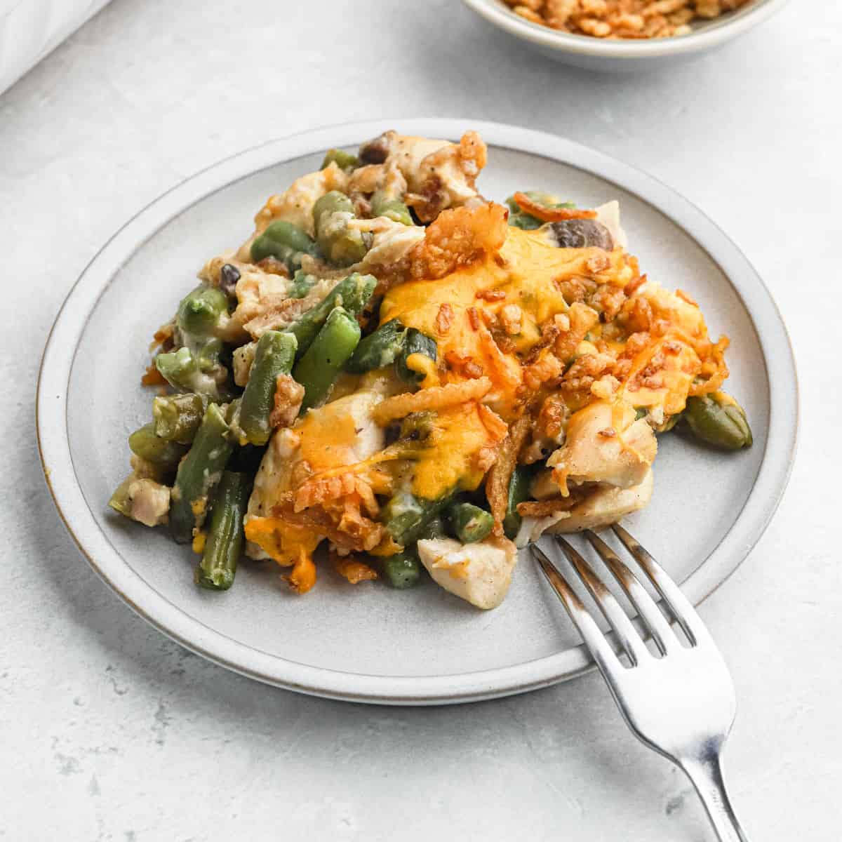 A plate of chicken green bean casserole topped with melted cheese and crispy onions, served with a fork on a light-colored plate and surface.