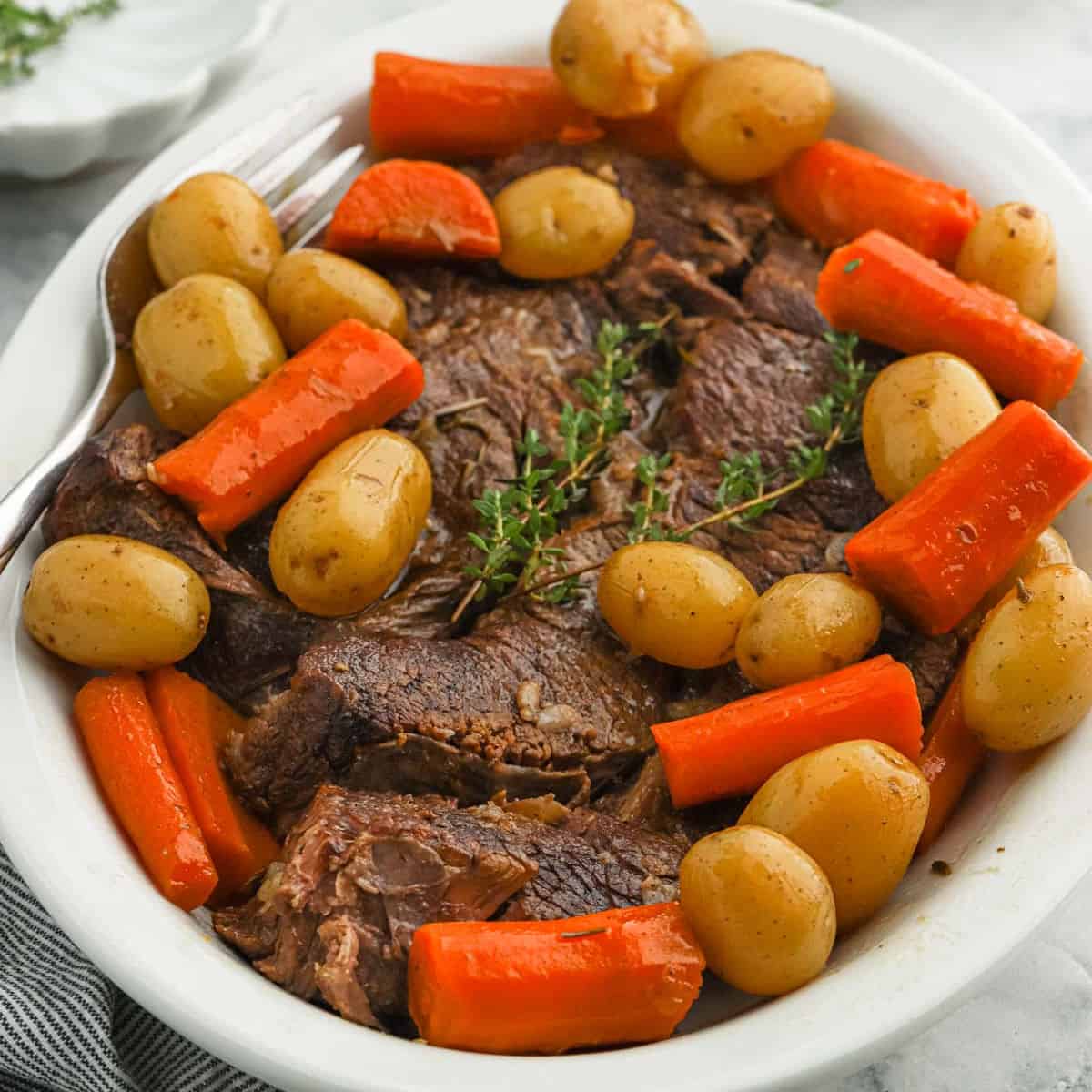 A white dish filled with Dutch oven pot roast, surrounded by whole baby potatoes, large carrot pieces, and garnished with sprigs of fresh thyme.