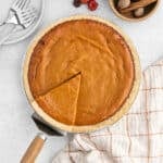 A top-down view of an Eagle Brand Pumpkin Pie with one slice being lifted out, alongside a checkered towel, a bowl of whole nutmeg and a grater, and a stack of plates with forks.