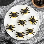 A white plate with seven Halloween Deviled Eggs, each topped with black olives arranged to look like spiders, sits on a black netted tablecloth. A bowl of sliced olives is nearby.