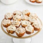 A glass cake stand displays a dozen mini cinnamon rolls topped with white icing, arranged in neat rows. In the background, a white plate holds extra mini cinnamon rolls for sharing.