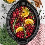 A Slow Cooker Christmas Potpourri filled with water, fresh cranberries, orange slices, rosemary sprigs, and cinnamon sticks sits surrounded by festive spices and greenery on a white surface.