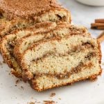 Close-up of sliced Snickerdoodle Bread, featuring a golden-brown crust and visible layers of cinnamon sugar filling inside. Crumbs are scattered on a white surface beside the loaf.