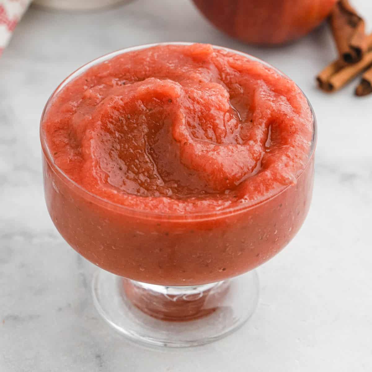 A glass dessert dish filled with thick, smooth reddish-brown strawberry applesauce sits on a light marble surface. Cinnamon sticks and part of an apple are blurred in the background.