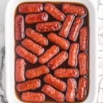 Cocktail sausages, reminiscent of classic Beanie Weenies, are coated in a glossy brown sauce and arranged neatly in a white rectangular dish. The dish sits on a marble surface next to a white and black striped towel.