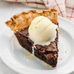A slice of brownie pie topped with a scoop of vanilla ice cream sits on a white plate, with a red and white checkered cloth in the background.