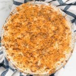 A round Butterfinger Pie with a creamy filling, topped with a generous layer of crumbled cookies, sits on a striped blue and white cloth. The pie is in a foil pan, viewed from above.