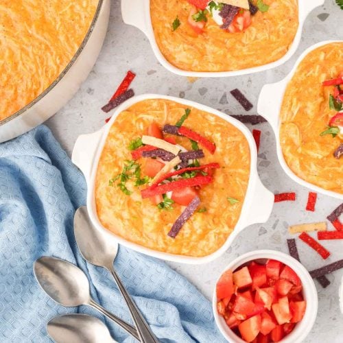 Bowls of creamy chicken tortilla soup, inspired by Chili's chicken enchilada soup recipe, topped with shredded cheese, crispy tortilla strips, and chopped tomatoes, arranged on a countertop with a blue towel, spoons, and diced tomatoes.