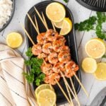 Grilled shrimp skewers, marinated in a flavorful grilled shrimp marinade, are garnished with parsley on a black platter and surrounded by lemon slices. A bowl of white rice and a striped napkin sit nearby on a white wooden surface.