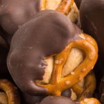 A close-up of crunchy Peanut Butter Pretzel Bites partially dipped in smooth, dark chocolate. The golden-brown pretzels with visible salt grains contrast perfectly with the glossy chocolate coating.
