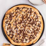 A freshly baked s'mores pizza topped with toasted mini marshmallows, melted chocolate, and chocolate chips on a round baking tray, served alongside plates and a stack of graham crackers.
