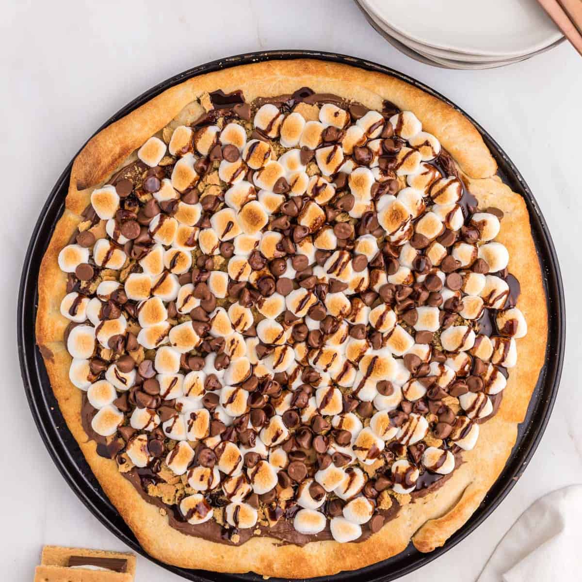 A freshly baked s'mores pizza topped with toasted mini marshmallows, melted chocolate, and chocolate chips on a round baking tray, served alongside plates and a stack of graham crackers.
