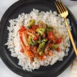 A black plate with white rice topped with sautéed teriyaki vegetables—broccoli, red bell pepper, onion, and mushroom—garnished with black sesame seeds. A gold fork is placed to the right of the plate.