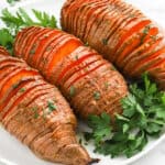 Three baked Hasselback Sweet Potatoes are sliced thinly and garnished with fresh parsley, served on a white plate with extra parsley sprinkled around for a vibrant finish.