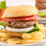 A hamburger with lettuce, onion, tomato, and a beef patty in a bun, served on a white plate with crinkle-cut potato chips—perfect inspiration for Blackstone ground beef recipes. Another burger is slightly blurred in the background.