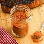 A jar filled with reddish-brown steak rub sits on a wooden surface next to a spoonful of the seasoning, a piece of grilled meat, and a red checkered cloth.