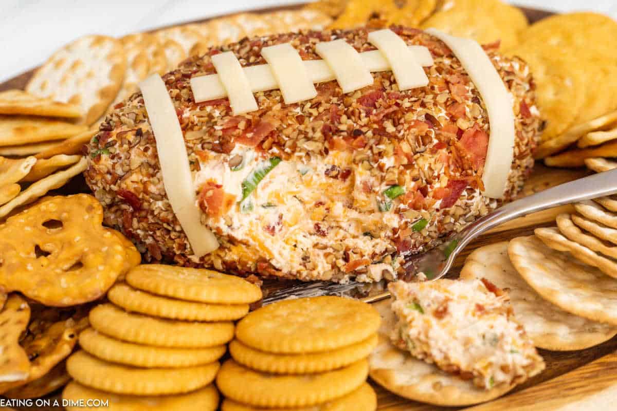 A Football Cheese Ball shaped like a football, coated with chopped nuts and topped with cheese strips as laces, is surrounded by round crackers and pretzels on a wooden tray. A spoon scoops out some of the delicious cheese ball.