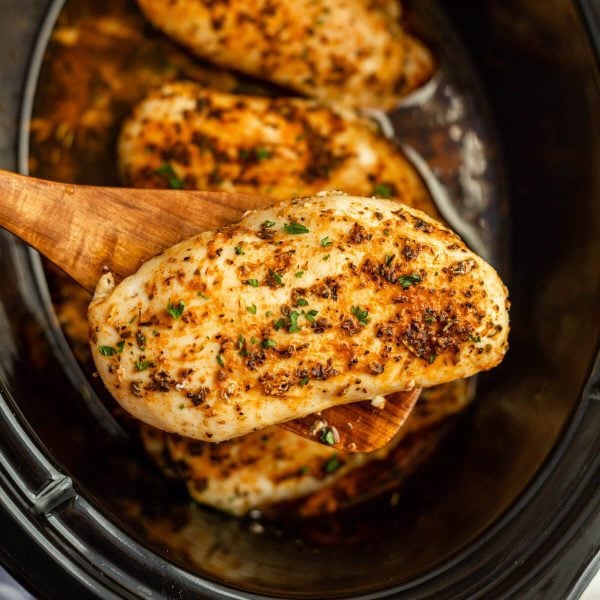 A wooden spoon lifts a juicy, seasoned slow cooker chicken breast from the pot, with more chicken breasts visible inside. The meat is garnished with herbs and looks perfectly cooked and tender.
