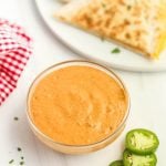 A glass bowl of creamy orange Taco Bell Creamy Jalapeño Sauce sits next to sliced jalapeños, with a plate of cheese quesadilla in the background and a red and white checkered cloth nearby.