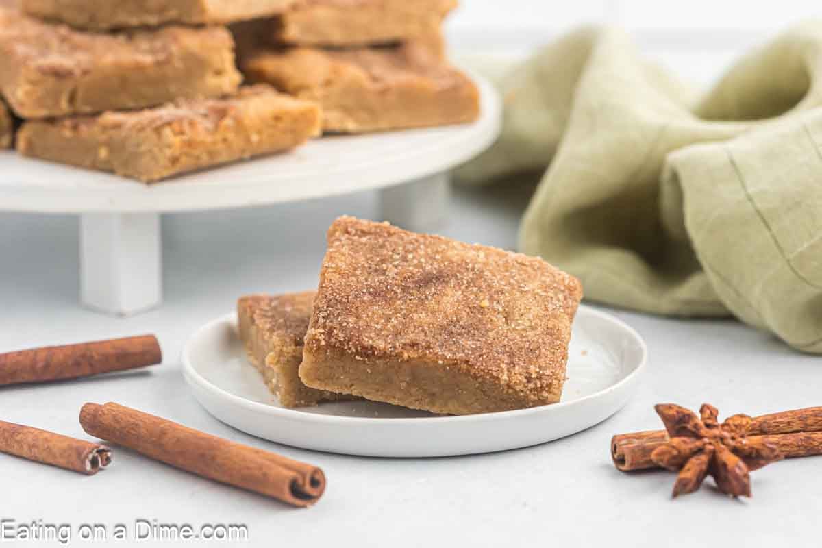 Two square snickerdoodle blondies stacked on a plate with cinnamon sticks laying nearby.