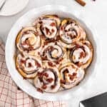A round white baking dish filled with Bacon Cinnamon Rolls, each topped with white icing and crumbled bacon pieces, sits on a white surface beside a red and white checkered towel.