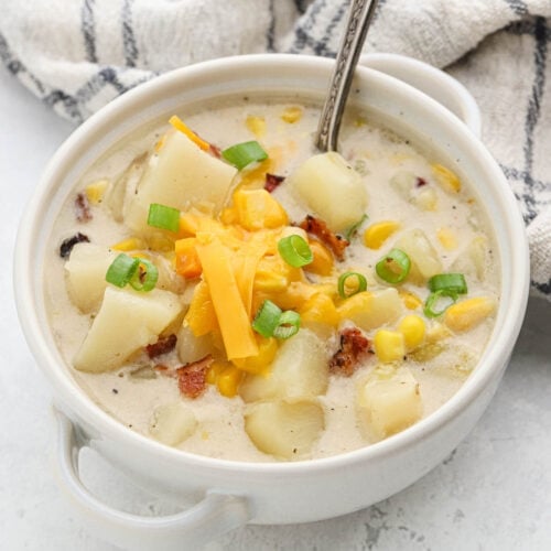 A bowl of Crockpot Potato Corn Chowder with corn, diced potatoes, bacon bits, shredded cheddar cheese, and chopped green onions, served with a spoon and a striped cloth in the background.