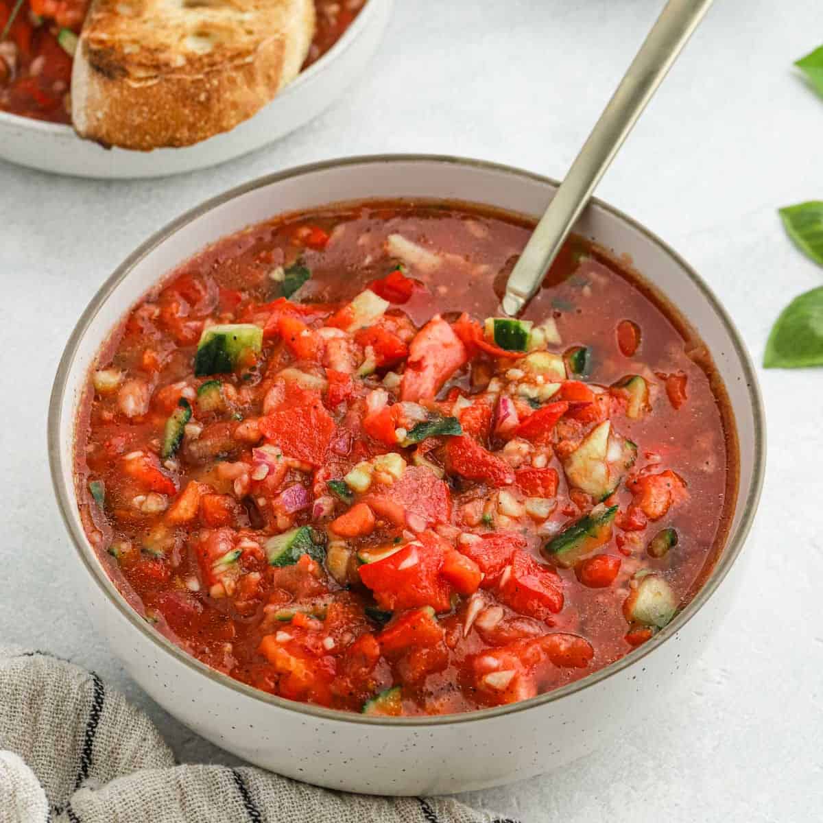 A bowl of chunky gazpacho soup with diced tomatoes, cucumbers, and peppers, served with a spoon. Inspired by the Ina Garten Gazpacho Recipe, a slice of toasted bread is visible in the background.