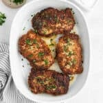 Four golden-brown, Parmesan Crusted Pork Chops garnished with chopped parsley are arranged in a white baking dish on a light surface, with a striped napkin partially visible beside the dish.