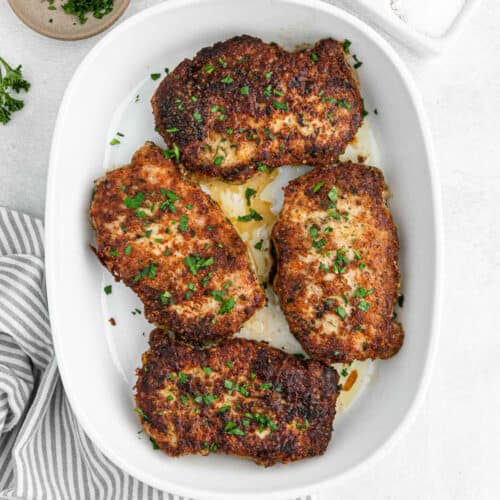 Four golden-brown, Parmesan Crusted Pork Chops garnished with chopped parsley are arranged in a white baking dish on a light surface, with a striped napkin partially visible beside the dish.