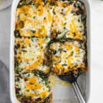 A casserole dish filled with a delicious Vegetarian Breakfast Casserole, topped with melted cheese. One large piece has been removed, revealing layers of veggies, cheese, and sauce. A serving spatula rests in the empty space.
