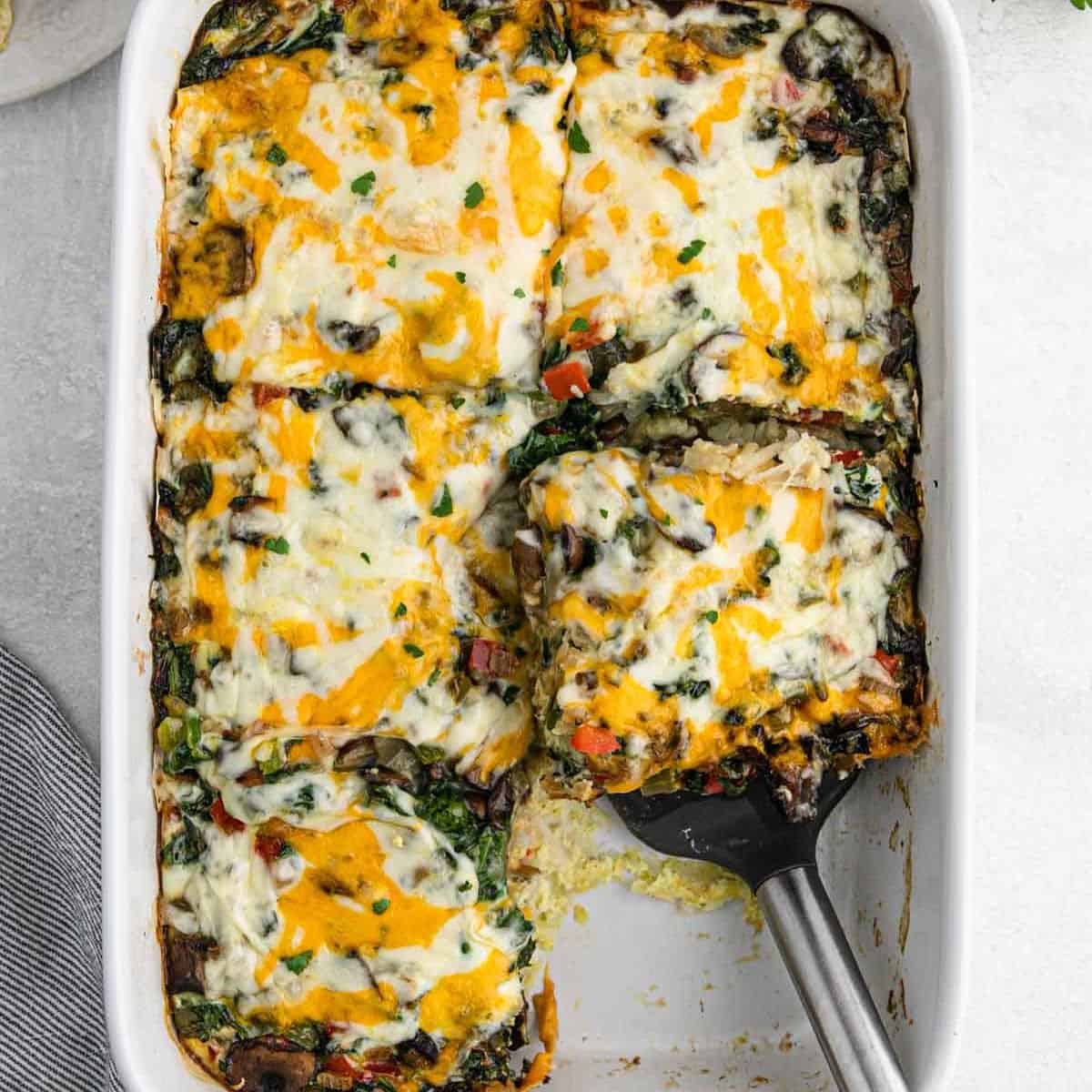 A casserole dish filled with a delicious Vegetarian Breakfast Casserole, topped with melted cheese. One large piece has been removed, revealing layers of veggies, cheese, and sauce. A serving spatula rests in the empty space.