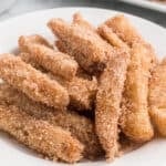 A plate filled with golden-brown churros and crispy apple fries, both coated in cinnamon sugar, arranged in a pile and ready to eat.