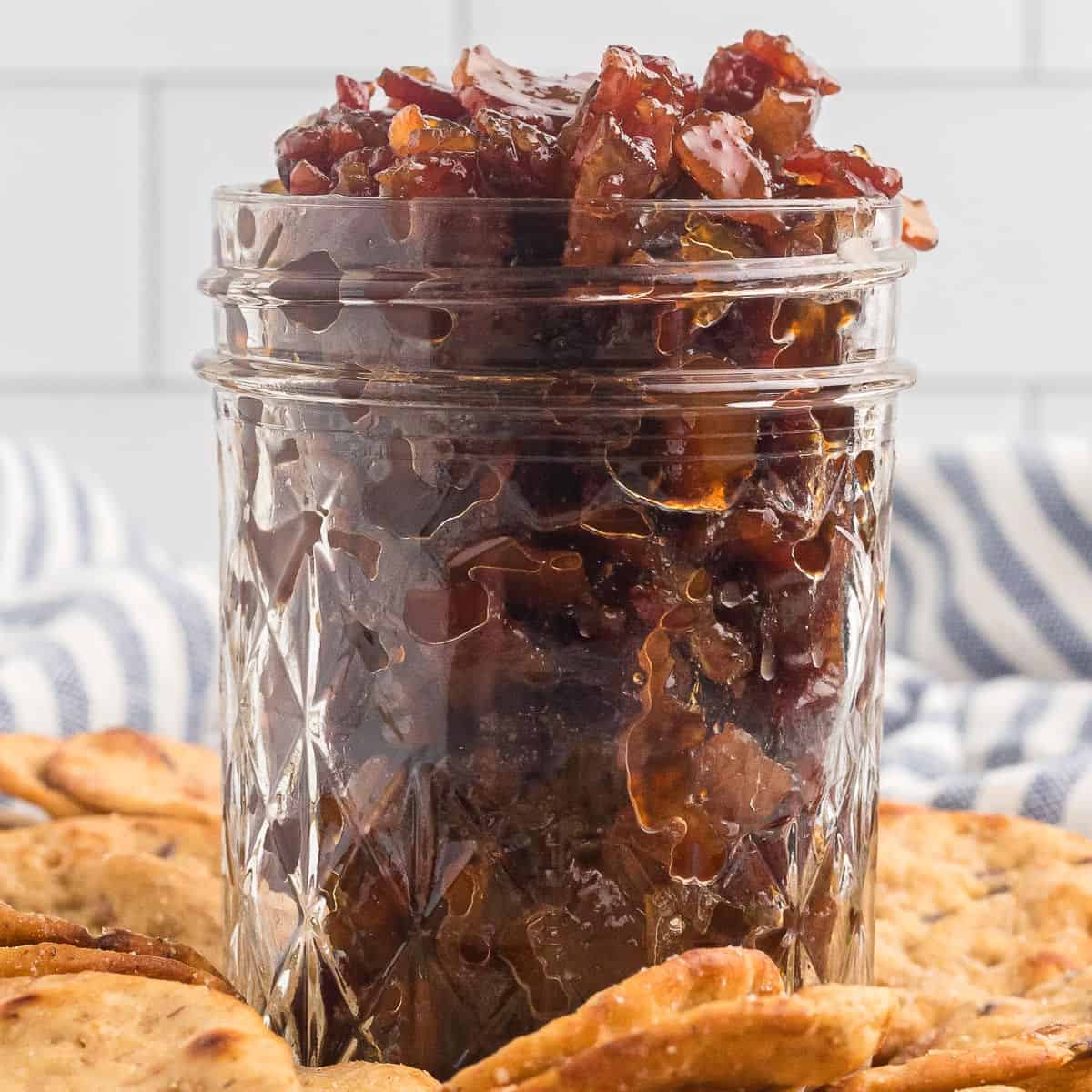 A glass jar filled with savory bacon jam sits on a plate surrounded by round crackers, with a blue and white striped towel in the background.
