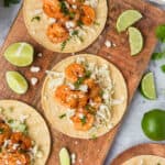 Three Blackened Shrimp Tacos on corn tortillas are arranged on a wooden board, topped with slaw, crumbled cheese, and herbs, with lime wedges scattered around for garnish.