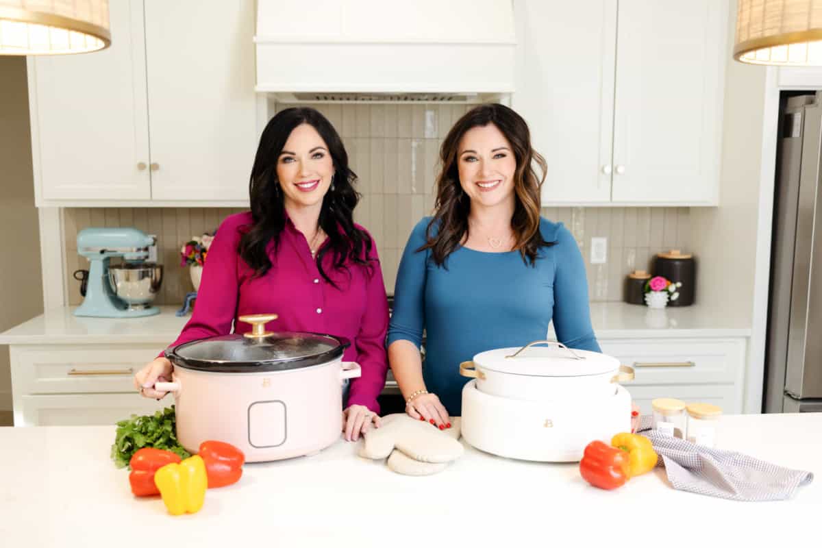 Carrie and Christina standing by two crock pots with bell peppers, fresh herbs, pot holders and a towel on the counter.