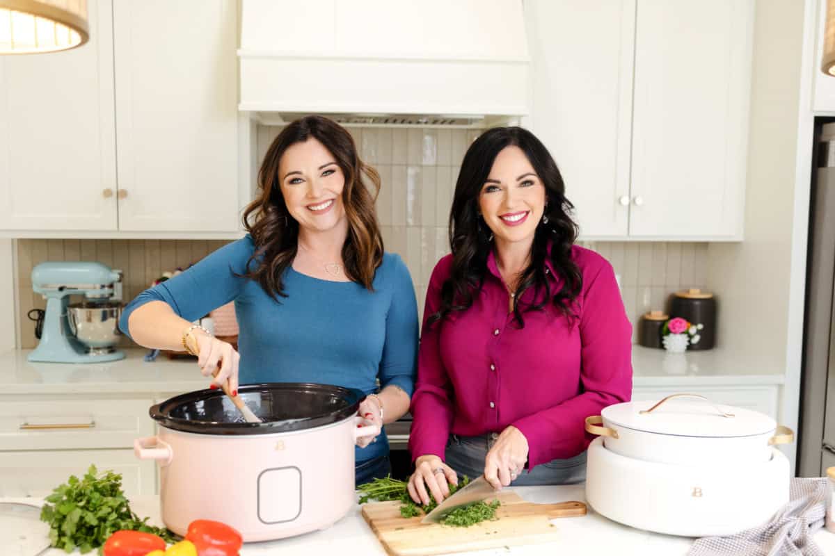 Christina stirring food in the crockpot while Carrie is chopping fresh herbs on a cutting board with a crock pot nearby on the counter.