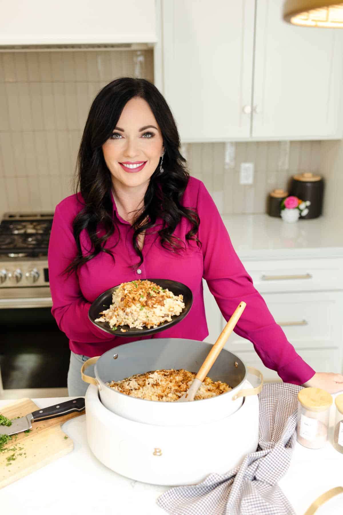 Carrie in a pink top holding a plate of food with a crock pot with food inside on the counter.