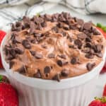 A white bowl filled with creamy Nutella dip topped with chocolate chips, surrounded by fresh strawberries; a striped cloth is in the background.