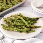 A white plate filled with Parmesan Roasted Green Beans, topped with grated cheese, sits on a white tiled surface next to a baking tray and stacked white plates with forks.